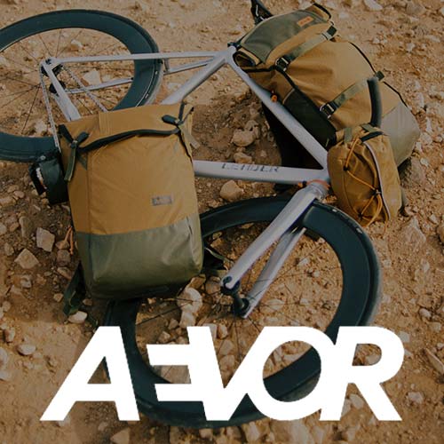 Ein weißes Fahrrad mit hellbraunen und grünen Taschen liegt auf felsigem Grund. Der fettgedruckte weiße Text AEVOR ist am unteren Rand des Bildes eingeblendet.