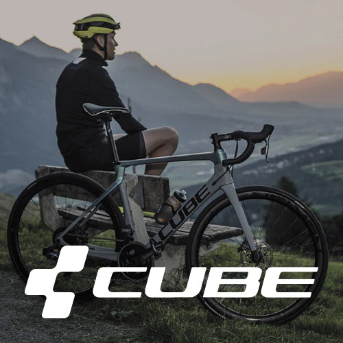 Ein Radfahrer mit Helm sitzt auf einer Bank neben einem Rennrad und blickt auf die Berge bei Sonnenuntergang. Das Logo der Marke Cube ist prominent im Vordergrund zu sehen.