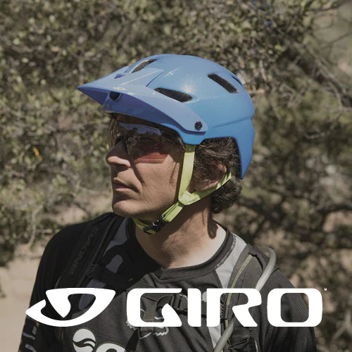 Ein Mann mit blauem Fahrradhelm und Sonnenbrille steht im Freien zwischen Bäumen, wobei das weiße GIRO-Logo am unteren Bildrand hervorgehoben wird.