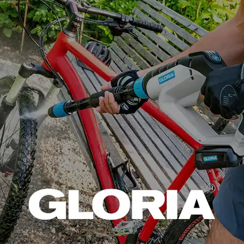 Eine Person verwendet einen Hochdruckreiniger der Marke GLORIA, um das Vorderrad eines roten Fahrrads im Freien neben einer Metallbank zu reinigen.